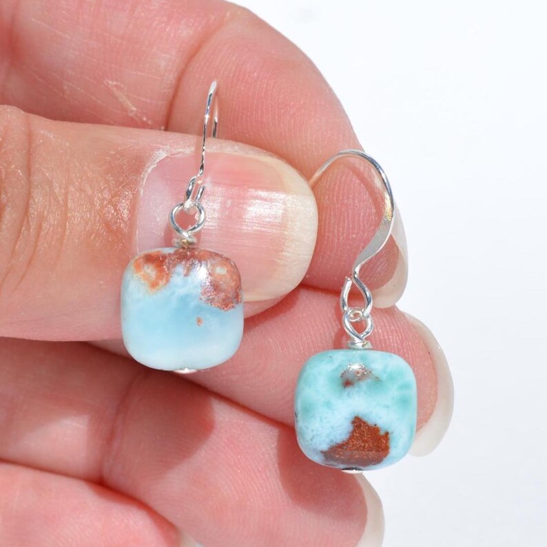 May include: A pair of square-shaped earrings with a light blue and brown pattern. The earrings are suspended from silver-colored hooks. The gemstones have a polished, smooth surface. The earrings are a type of jewelry.