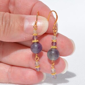 May include: A pair of dangle earrings with gold-colored hooks. Each earring features a large, round, purple bead, smaller purple and clear beads, and gold-colored accents. The earrings are displayed against a light background.