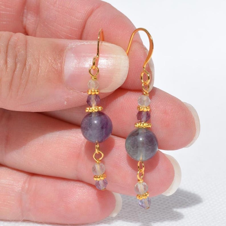 May include: A pair of dangle earrings with gold-colored hooks. Each earring features a large, round, purple bead, smaller purple and clear beads, and gold-colored accents. The earrings are displayed against a light background.