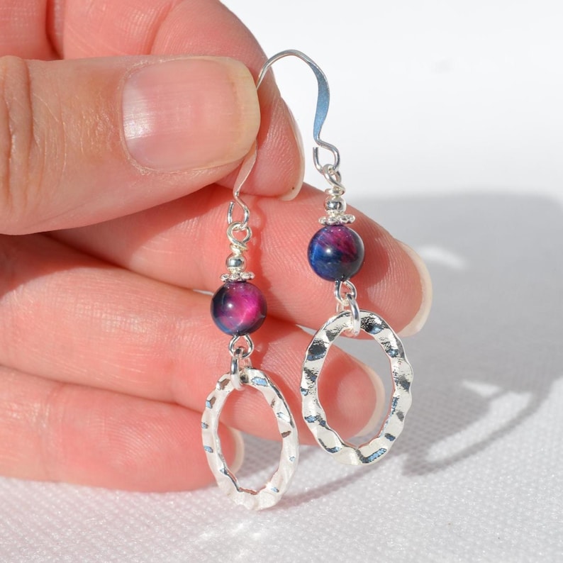 Op de afbeelding: Een paar hangende oorbellen met een zilverkleurige haak, een kleine ronde kraal met een dieppaars en blauw gemarmerd patroon, en een getextureerde, gehamerde zilverkleurige ovale ring aan de basis. De oorbellen zijn gemaakt van zilverkleurige metalen onderdelen.