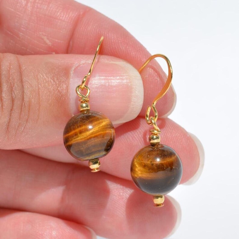 Puede incluir: Un par de pendientes colgantes con piedras preciosas de ojo de tigre redondas y pulidas, con bandas chatoyantes de color marr&oacute;n dorado. Cada piedra est&aacute; suspendida de un gancho de metal dorado y conectada por una peque&ntilde;a cuenta de metal dorado.