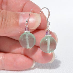 May include: A pair of dangle earrings with round, translucent green fluorite beads. Each earring has a small purple bead and silver-coloured ear wire. The earrings are held up against a light background.
