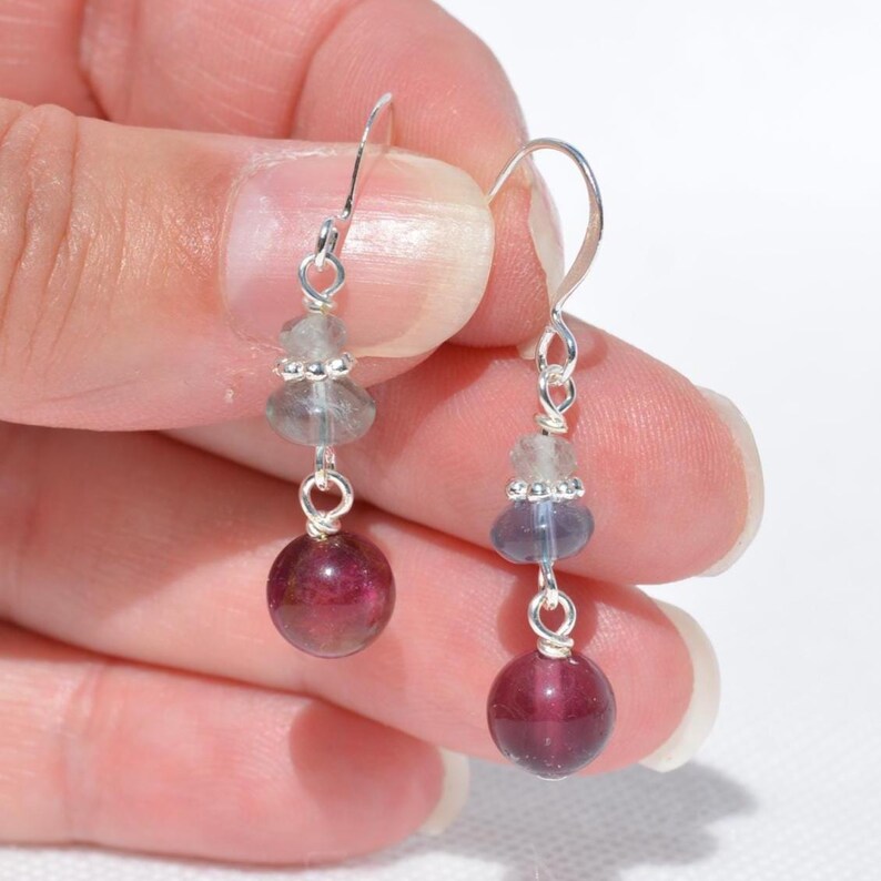 May include: A pair of dangle earrings with silver hooks. Each earring features a large, round, deep red bead at the bottom, with smaller, light blue and clear beads above. The earrings are held up by a silver hook.