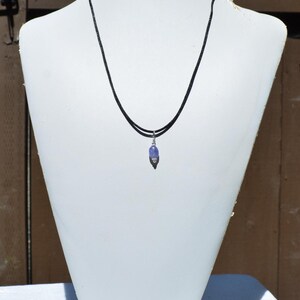 May include: A necklace with a small, light purple gemstone pendant. The pendant is suspended from a thin, black cord. The necklace is displayed on a white mannequin.