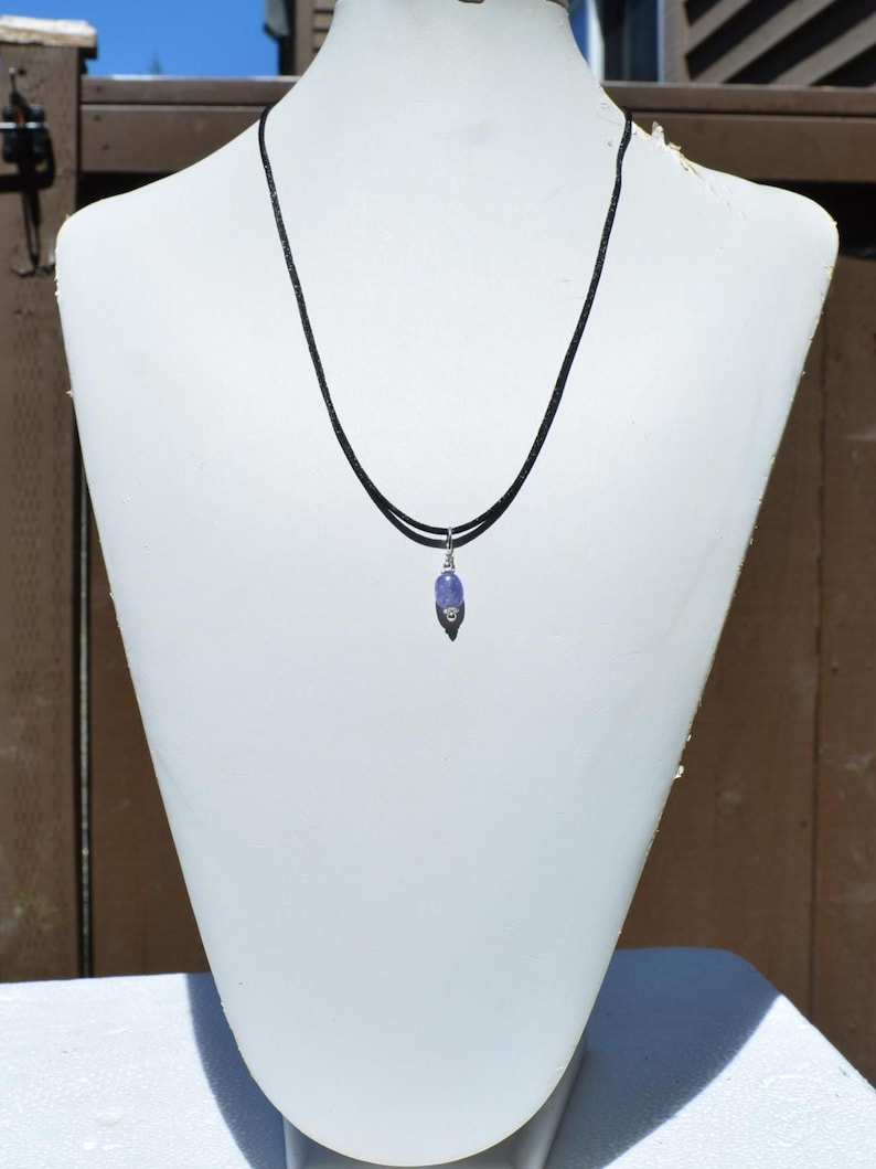 May include: A necklace with a small, light purple gemstone pendant. The pendant is suspended from a thin, black cord. The necklace is displayed on a white mannequin.