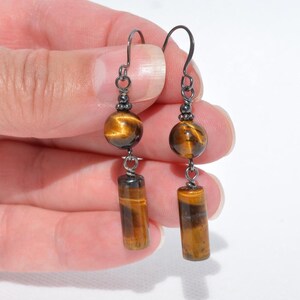May include: A pair of drop earrings featuring dark metal hooks, each with a round tiger's eye bead suspended above a cylindrical tiger's eye bead. The stones display characteristic golden-brown chatoyancy with dark bands, connected by small metal rings.