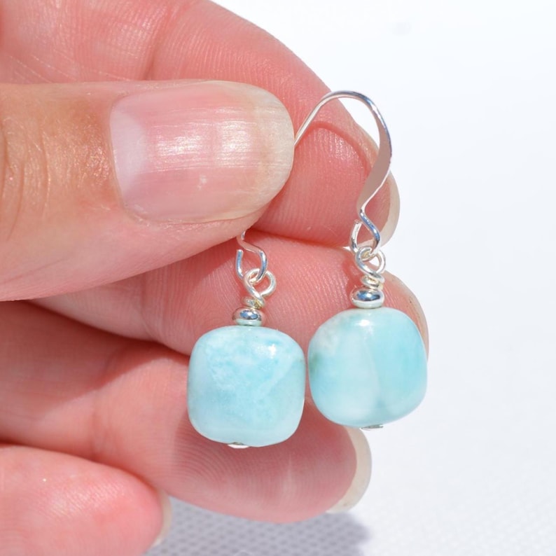May include: A pair of light blue, square-shaped earrings with silver-colored hooks. The earrings are held up by a hand against a white background. The earrings have a smooth, polished appearance.