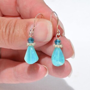 May include: A pair of drop earrings featuring light blue teardrop-shaped beads suspended from silver-toned hooks. Each earring includes a small, faceted dark blue bead and a silver-toned spacer bead positioned above the larger light blue drop.
