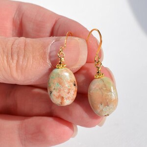May include: A pair of drop earrings featuring smooth, oval-shaped stones in shades of peach, tan, and light green. Each stone is suspended from a gold-toned metal hook with a small decorative bead at the top, connecting the stone to the wire.
