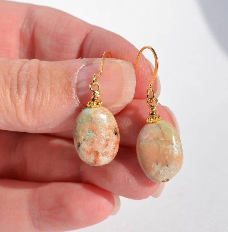 May include: A pair of drop earrings featuring smooth, oval-shaped stones in shades of peach, tan, and light green. Each stone is suspended from a gold-toned metal hook with a small decorative bead at the top, connecting the stone to the wire.