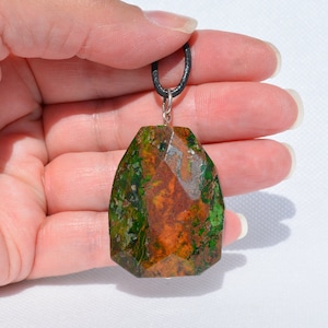 May include: A green and orange gemstone pendant with a faceted, irregular shape. The pendant is attached to a black cord.
