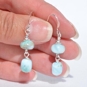 May include: A pair of dangle earrings featuring light blue and white gemstone beads. The earrings have a silver-colored hook and wire design. The beads are oval and round, with a polished finish. The earrings are held in a hand.