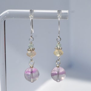 May include: Dangle earrings with a silver hook and a combination of beads. The earrings feature a large, round, translucent purple and white bead at the bottom, with smaller yellow and green beads above. The earrings are displayed on a clear stand.