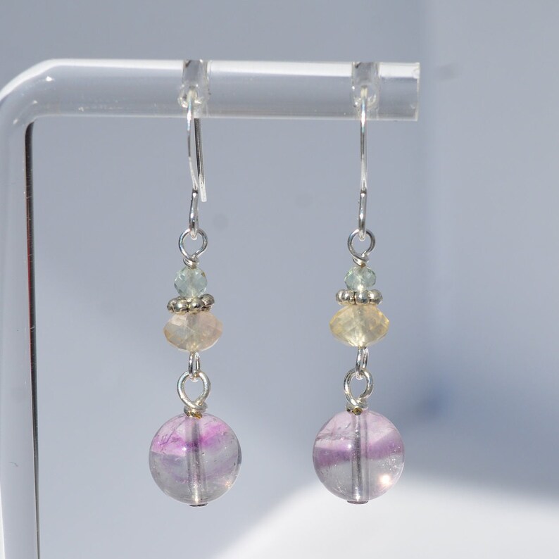 May include: Dangle earrings with a silver hook and a combination of beads. The earrings feature a large, round, translucent purple and white bead at the bottom, with smaller yellow and green beads above. The earrings are displayed on a clear stand.