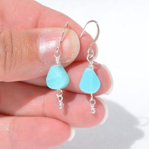 May include: A pair of drop earrings featuring light blue, teardrop-shaped beads suspended from silver-toned metal hooks. Each earring includes a small, clear accent bead at the bottom, connected by silver wire links to the main blue stone.
