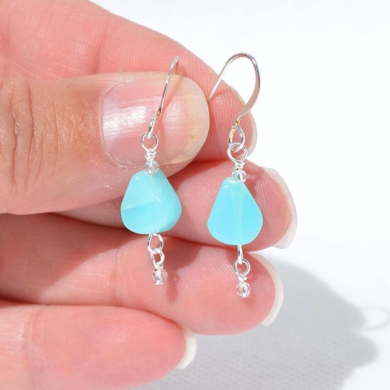 May include: A pair of drop earrings featuring light blue, teardrop-shaped beads suspended from silver-toned metal hooks. Each earring includes a small, clear accent bead at the bottom, connected by silver wire links to the main blue stone.