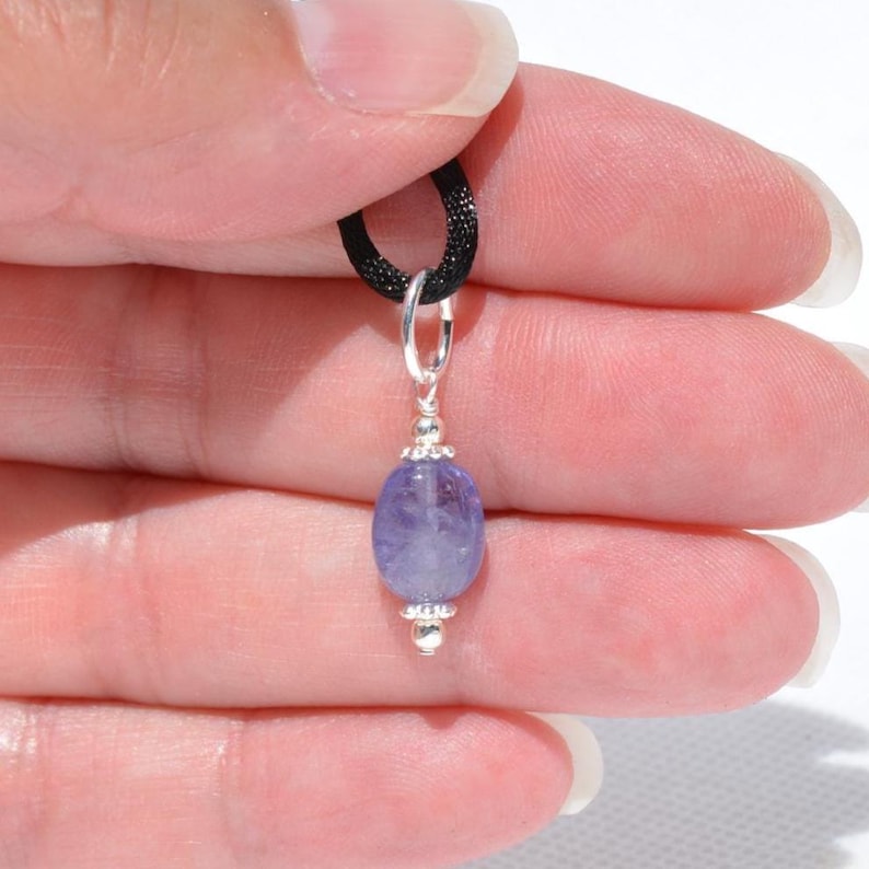 May include: A small, oval-shaped pendant with a light purple gemstone. The gemstone is suspended from a silver-colored bail and a black cord. The pendant is held in a person's hand.