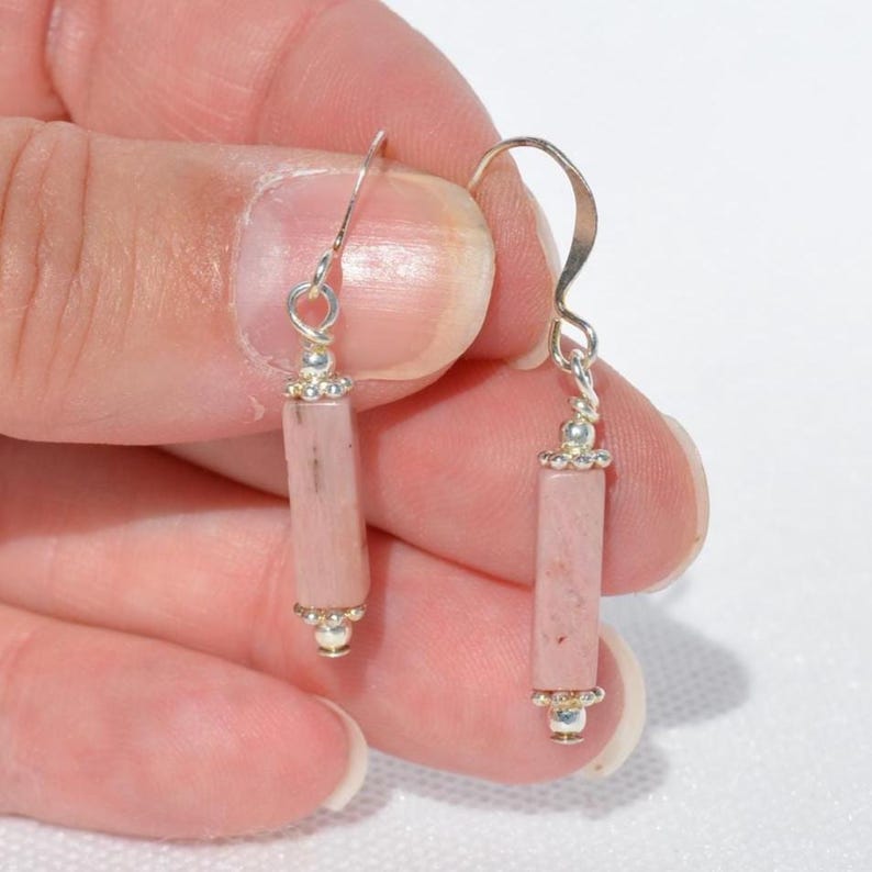 May include: A pair of drop earrings featuring cylindrical pink stone beads suspended from silver-toned metal hooks. Each earring includes a small silver-toned bead at the top and bottom of the stone cylinder, creating a simple and balanced design.