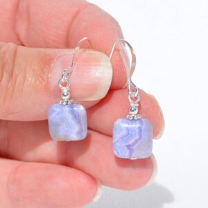 May include: A pair of silver-tone earrings featuring square, light blue gemstones with a marbled pattern. The earrings have a simple hook design and small decorative silver beads.