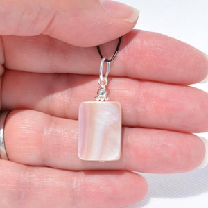 May include: A rectangular pendant with a pearlescent, pinkish-white surface. The pendant is attached to a black cord with a silver-colored bail. The pendant is held in a hand.