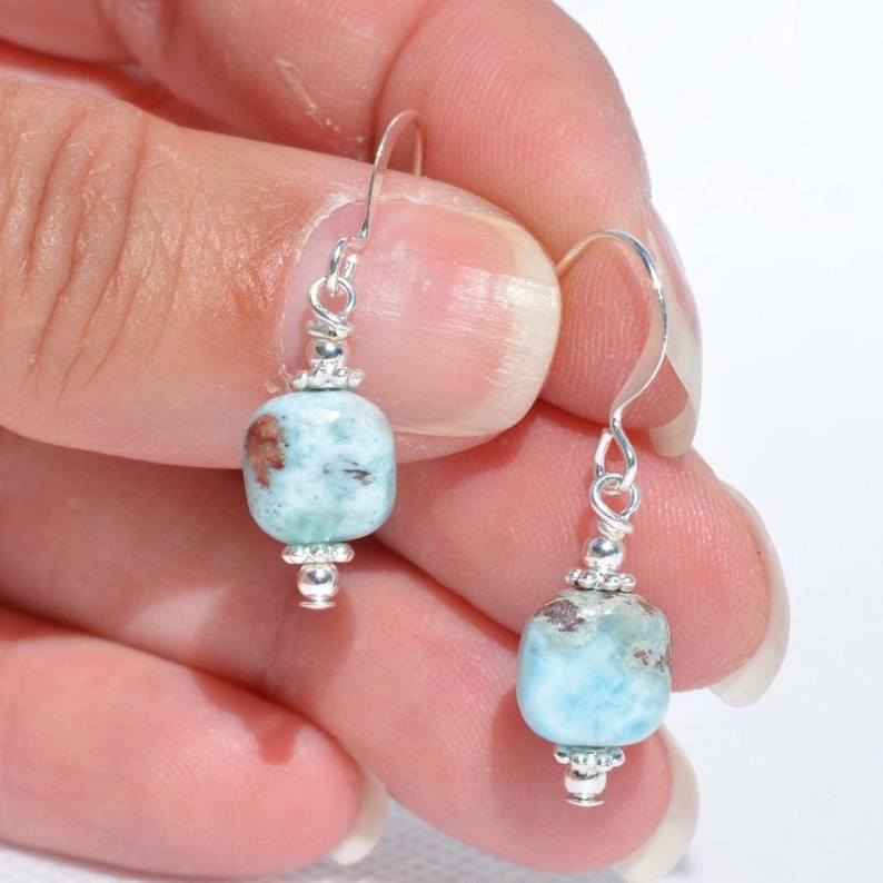 May include: A pair of dangle earrings featuring light blue and brown gemstone beads. The beads are rectangular with rounded edges and are suspended from silver-colored hooks. The earrings are held in a person's hand against a white background.