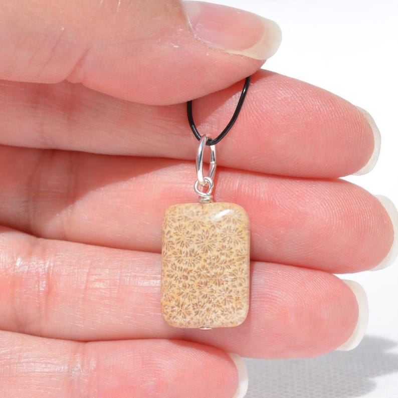 Op de afbeelding: Een rechthoekige hanger van beige fossiel koraalsteen met een gedetailleerd bloemachtig patroon. De steen is bevestigd met een zilverkleurig metalen oogje en hangt aan een dun zwart koord, wat zorgt voor een eenvoudig ontwerp van de ketting.