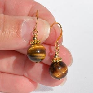 Puede incluir: Un par de pendientes colgantes con piedras preciosas de ojo de tigre redondas y pulidas con bandas chatoyantes marrones y doradas. Cada piedra cuelga de un gancho de metal dorado mediante una peque&ntilde;a tapa de cuentas con borde texturizado.