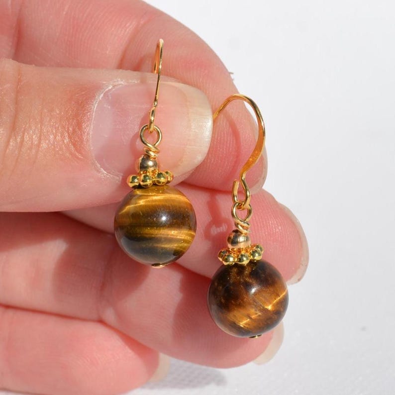 Puede incluir: Un par de pendientes colgantes con piedras preciosas de ojo de tigre redondas y pulidas con bandas chatoyantes marrones y doradas. Cada piedra cuelga de un gancho de metal dorado mediante una peque&ntilde;a tapa de cuentas con borde texturizado.