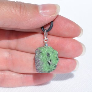 May include: A green and pink raw gemstone pendant on a black cord. The pendant is attached to a silver bail.