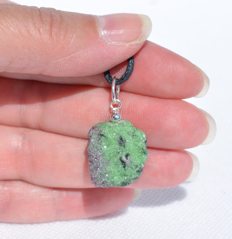 May include: A green and pink raw gemstone pendant on a black cord. The pendant is attached to a silver bail.