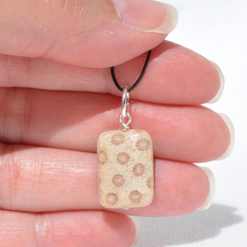Op de afbeelding: Een rechthoekige hanger van beige gefossiliseerd koraal met een natuurlijk cirkelvormig patroon over de hele steen. De hanger is bevestigd aan een zilverkleurig metalen oogje en hangt aan een dun zwart koord met een gladde afwerking.