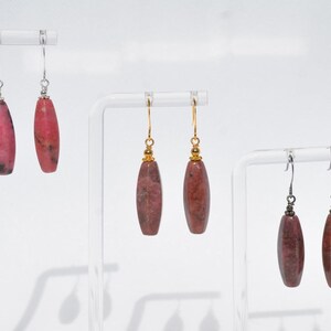 May include: Three pairs of drop earrings featuring elongated, cylindrical stone beads in varying shades of pink and reddish-brown. Each pair hangs from metal hooks, with the first pair on silver, the second on gold, and the third on dark metal.