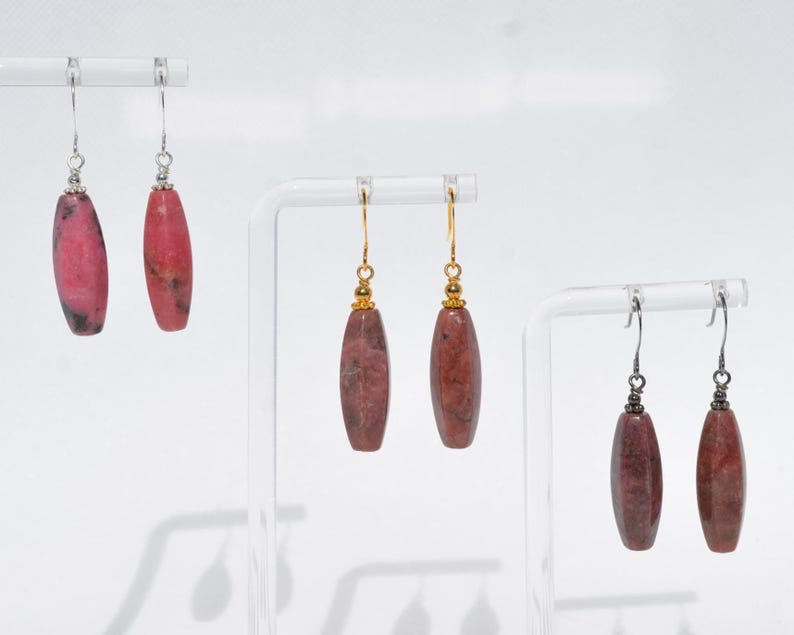 May include: Three pairs of drop earrings featuring elongated, cylindrical stone beads in varying shades of pink and reddish-brown. Each pair hangs from metal hooks, with the first pair on silver, the second on gold, and the third on dark metal.