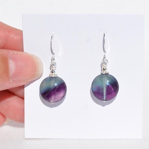 May include: A pair of drop earrings with spherical fluorite beads, displaying purple and teal hues. The beads are attached to silver-coloured hooks with decorative silver detailing. The earrings are presented against a white backdrop.