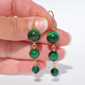 May include: A pair of gold-colored drop earrings featuring three green, spherical beads. The beads are connected by small gold-colored loops. The earrings are displayed against a white background.