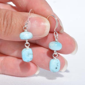 May include: A pair of dangle earrings featuring two light blue, oval-shaped stones connected by silver-colored wire. The earrings have a simple hook design. The stones have a slightly speckled appearance.