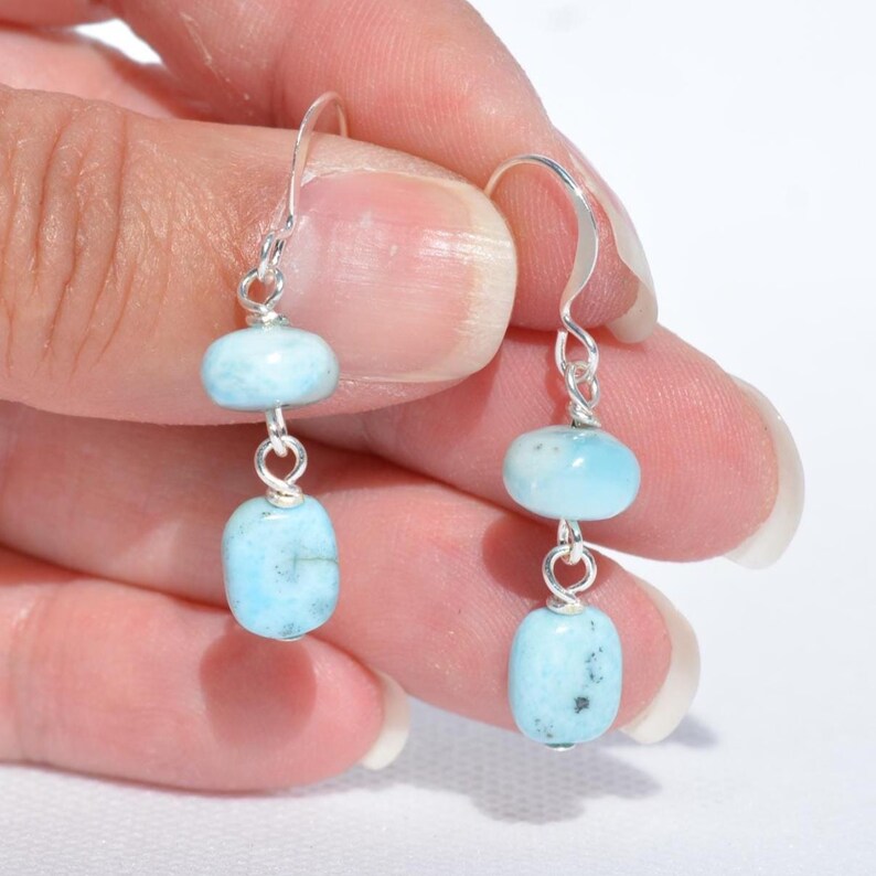 May include: A pair of dangle earrings featuring two light blue, oval-shaped stones connected by silver-colored wire. The earrings have a simple hook design. The stones have a slightly speckled appearance.