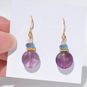 May include: Dangle earrings with a large, round, purple fluorite bead. The earrings have gold-coloured hooks and accents, with a small blue bead and a clear bead above the purple bead. The earrings are displayed on a white background.