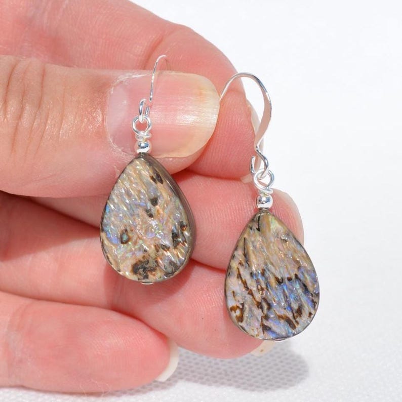 May include: A pair of teardrop-shaped earrings with a silver hook. The earrings have a marbled pattern in shades of brown, beige, and blue. The earrings are held up by a person's hand.