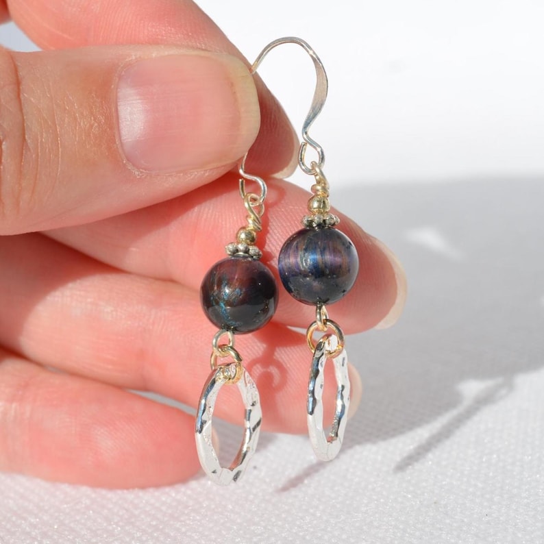 Op de afbeelding: Hangende oorbellen met donkerblauwe en paarse iriserende bolvormige kralen, geaccentueerd met kleine zilverkleurige metalen kraaltjes. Elke oorbel eindigt in een getextureerde, gehamerde zilverkleurige ovale ring die aan een haakje hangt.