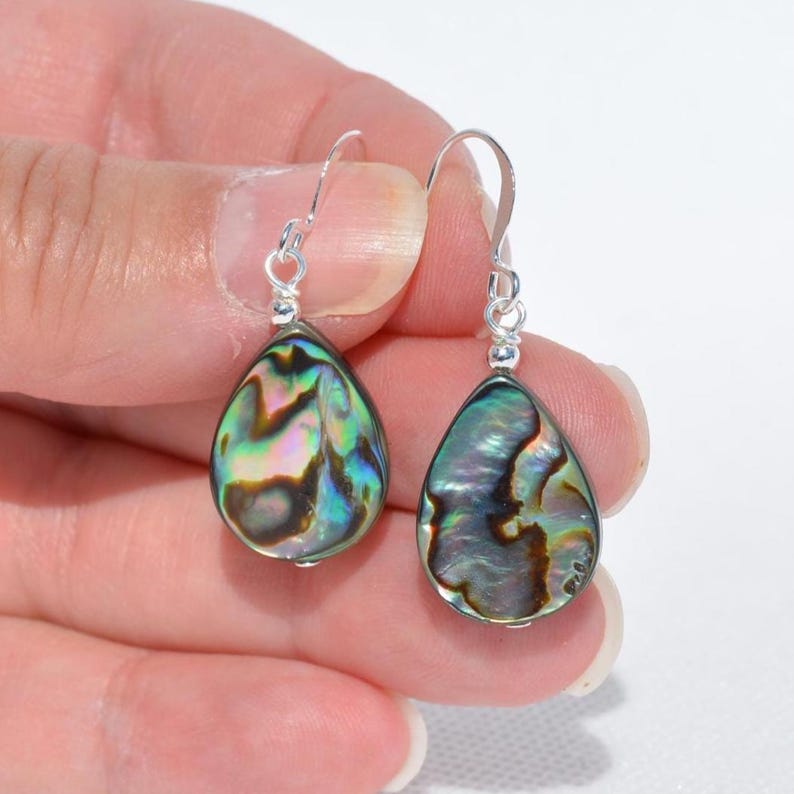 May include: A pair of teardrop-shaped earrings with iridescent abalone shells. The earrings have silver-colored hooks. The abalone shells display a mix of green, blue, purple, and brown colors. The earrings are displayed against a white background.