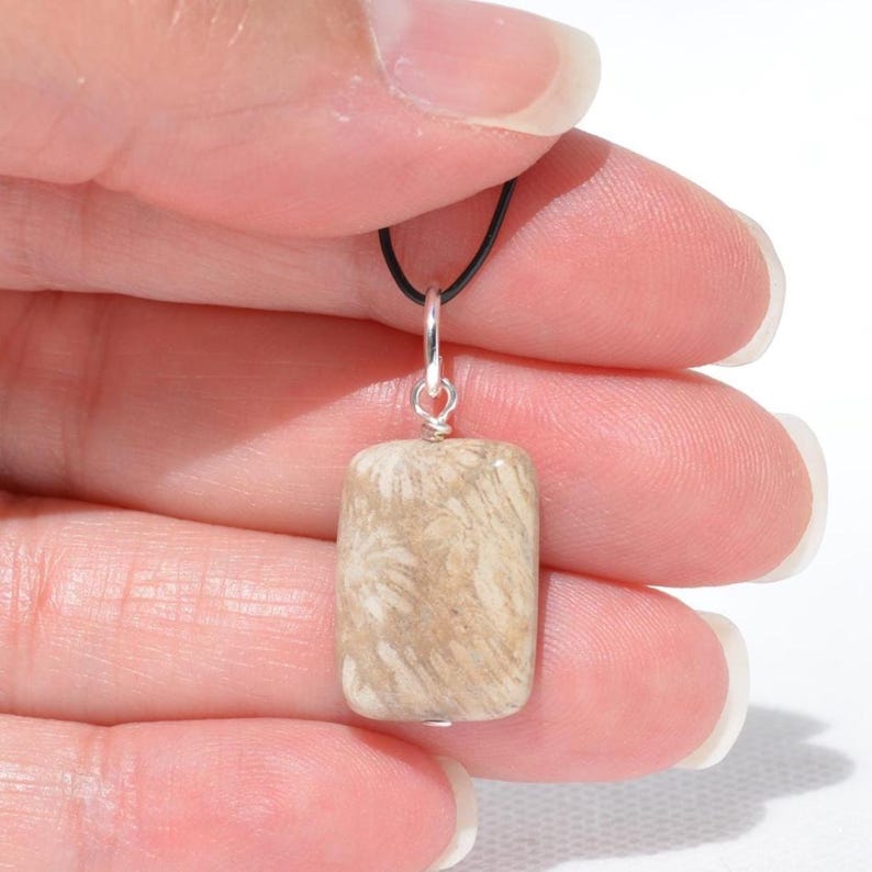 Op de afbeelding: Een rechthoekige hanger met een beige en cr&egrave;mekleurige gefossiliseerde koraalsteen met een opvallend stervormig patroon. De steen hangt aan een zilverkleurig metalen oogje dat is bevestigd aan een dun zwart koord voor een eenvoudig collier.