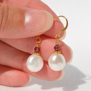 Puede incluir: Pendientes de oro con una perla blanca y detalles de piedras preciosas rojas.