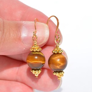 Puede incluir: Un par de pendientes colgantes con piedras preciosas de ojo de tigre redondas y pulidas en tonos marr&oacute;n dorado. Cada piedra est&aacute; decorada con peque&ntilde;as cuentas de metal dorado texturizado en la parte superior e inferior, suspendidas de ganchos.
