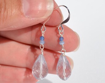 Op de afbeelding: Een paar bungelende oorbellen met gefacetteerde, druppelvormige lichtblauwe kristallen die aan zilverkleurige metalen haken hangen. Elke oorbel bevat twee kleine, ronde blauwe kralen boven de kristallen druppel, verbonden door zilverkleurige draadschakels.