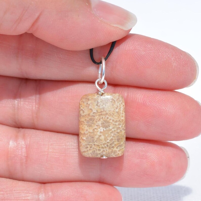 Op de afbeelding: Een rechthoekige hanger van beige gefossiliseerd koraal met een poreus, getextureerd oppervlak. De steen is bevestigd aan een zilverkleurige metalen ring en hangt aan een dun zwart koord, met natuurlijke beige en lichtbruine kleurnuances.