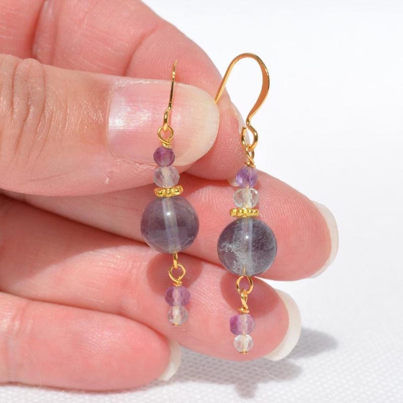 May include: A pair of gold-colored dangle earrings. Each earring features a large, round, purple bead, smaller purple and clear beads, and gold-colored accents. The earrings are held up by gold-colored hooks.