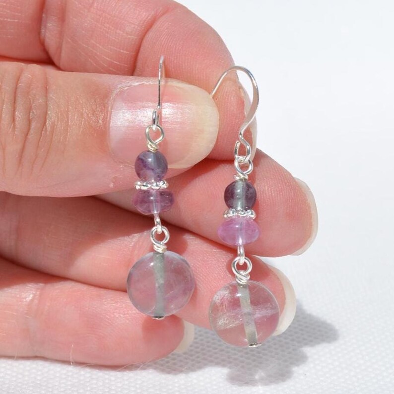 May include: Dangle earrings with purple and pink beads. The earrings have a silver hook and are made up of several beads, including a large, round, translucent pink bead at the bottom. The earrings are held up by a hand.