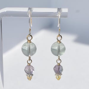 May include: A pair of dangle earrings featuring a light green, round bead above a faceted, purple bead. The earrings have silver hooks and gold-colored accents. A small, yellow bead is at the bottom of each earring.