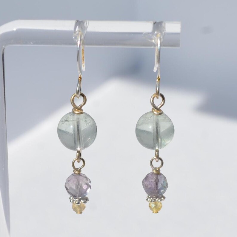May include: A pair of dangle earrings featuring a light green, round bead above a faceted, purple bead. The earrings have silver hooks and gold-colored accents. A small, yellow bead is at the bottom of each earring.